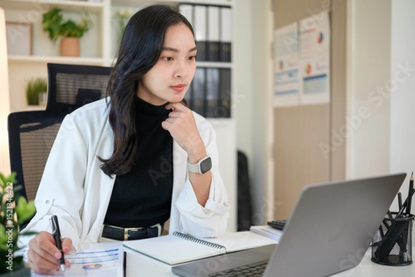 Obraz Focused businesswoman reviewing financial data and using laptop in a modern office.