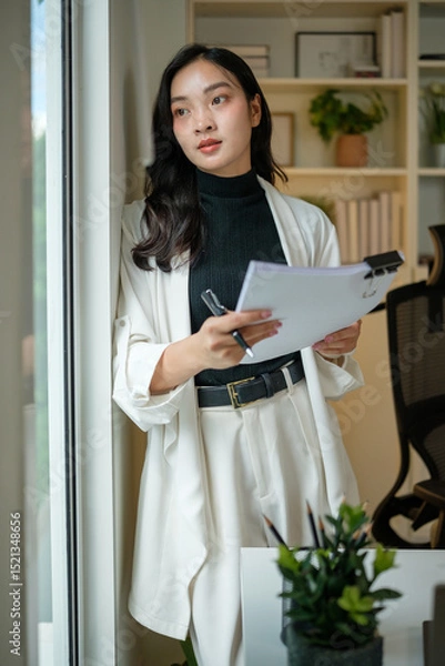 Obraz Thoughtful professional businesswoman in smart casual attire planning her tasks near a bright window.