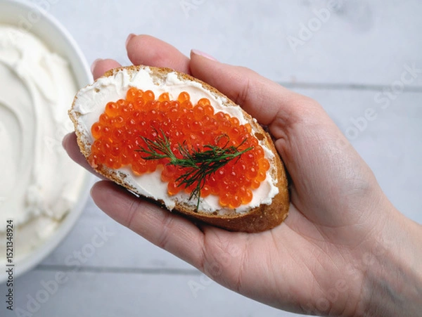 Obraz A hand presents a piece of bread adorned with cream cheese and caviar, highlighting a luxurious culinary treat.
