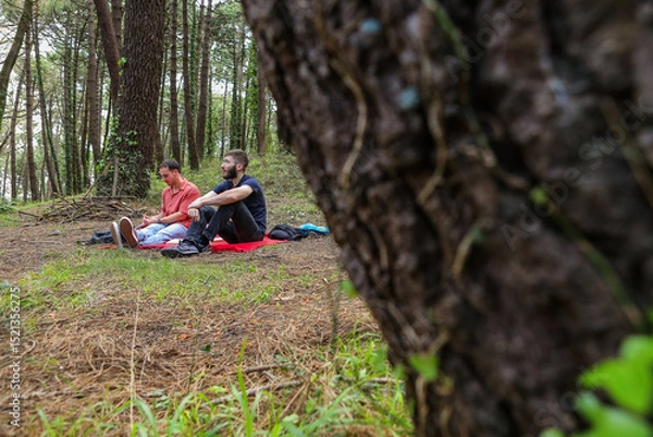 Obraz Two men in the forest resting and enjoying nature. Lying on a red blanket, reading, eating, and chatting