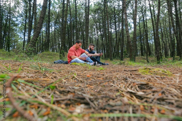 Obraz Two men in the forest resting and enjoying nature. Lying on a red blanket, reading, eating, and chatting