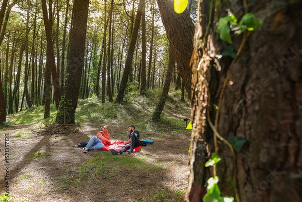 Obraz Two men in the forest resting and enjoying nature. Lying on a red blanket, reading, eating, and chatting
