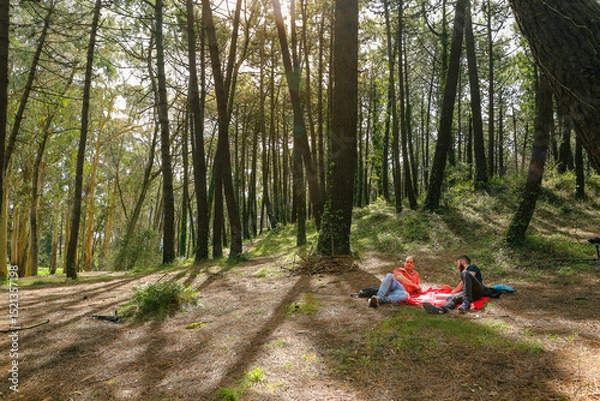 Obraz Two men in the forest resting and enjoying nature. Lying on a red blanket, reading, eating, and chatting