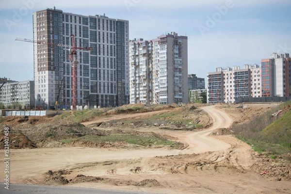 Fototapeta construction of the foundation of a new residential building using a tower crane in a large neighborhood with new tall modern multi-storey buildings on the background of which a winding dirt road lead