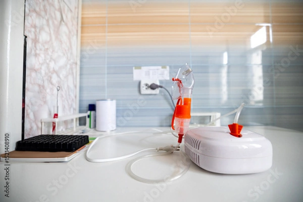 Fototapeta A close-up view shows a person's hands holding an inhaler device with an orange mask. The device is connected to a white compressor, all resting on a white table in a medical setting with tiled walls