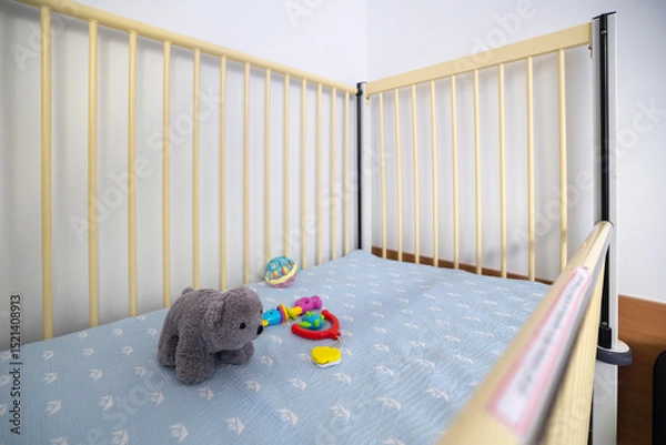 Fototapeta An empty hospital bed, designed for infants or young children, features light rails, a patterned mattress, and toys, set in a sterile room.