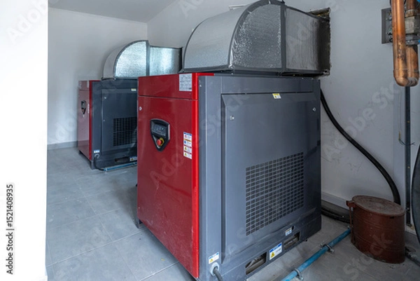 Fototapeta Two large industrial screw compressors, capable of 3220 liters per minute, stand in a utility room with insulated ductwork overhead, showcasing industrial machinery.