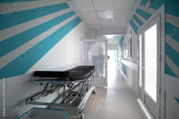 Fototapeta A hospital admission department corridor features striking blue and white diagonal stripes on the walls, with an empty stretcher and open doors leading further into the facility.