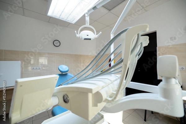 Fototapeta A modern dental clinic room features a blue dental chair, white dental unit with instruments, an overhead operating light, and a wall clock in a clean setting.