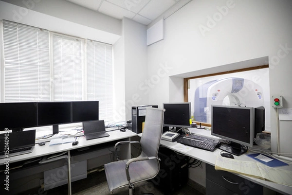Fototapeta A medical diagnostic control room features multiple computer monitors, laptops, and an office chair, with a window view into an adjacent CT scanner room on the basement floor of the facility.