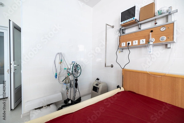 Fototapeta An intensive care ward for respiratory diseases features a red hospital bed, a lung ventilator, an oxygen concentrator, and an oxygen hub panel on a white wall.