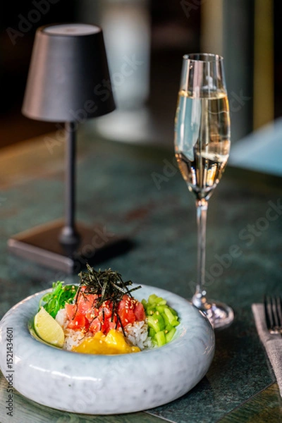 Fototapeta A gourmet poke bowl with fresh diced tuna, seaweed, cucumber, mango, and rice, garnished with sesame seeds and a lime wedge, served alongside a glass of champagne on a stylish table setting