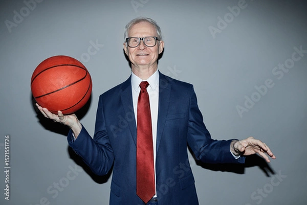 Fototapeta Portrait of senior Caucasian man wearing suit and tie smiling at camera while holding basketball against plain grey background in studio, camera flash