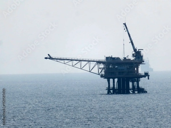 Obraz Offshore oil platform dominates the image, situated in a calm ocean under a hazy sky. A distant ship is visible. The background is a solid, hazy blue. The platform appears still, 