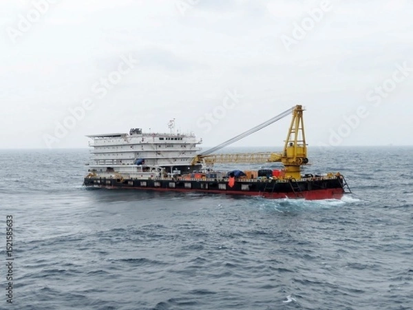 Obraz A large barge with a yellow crane sails across a dark blue ocean under a pale gray sky. The vessel appears sturdy and functional, navigating choppy waters. Its a scene of maritime industry at work.