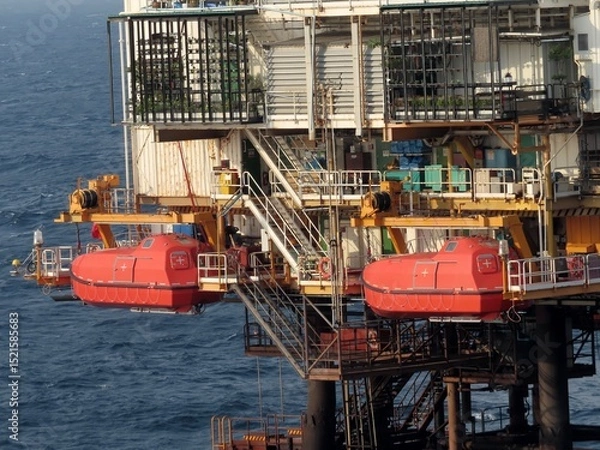 Obraz Two red life rafts hang from an offshore oil platform above a dark blue, choppy ocean. The platform shows signs of rust and wear. The overall atmosphere is one of industrial activity.