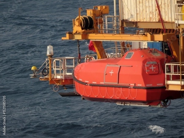 Obraz A vibrant orange lifeboat hangs suspended over a dark, choppy ocean. The vessel is attached to a yellow crane, ready for deployment. A sense of maritime safety and preparedness permeates the scene.