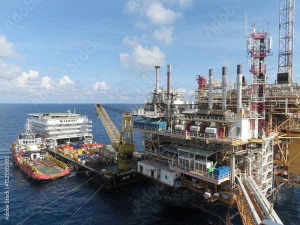 Obraz A large offshore oil platform dominates the image, with a support vessel alongside. The ocean is a deep blue, the sky is bright. The platform shows intricate structures, conveyors and equipment. 