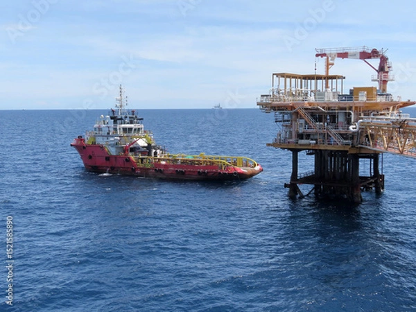 Obraz A red tugboat approaches a large offshore oil platform under a clear blue sky. The calm ocean reflects the sunlight, creating a serene yet industrial scene. Distant vessels dot the horizon.