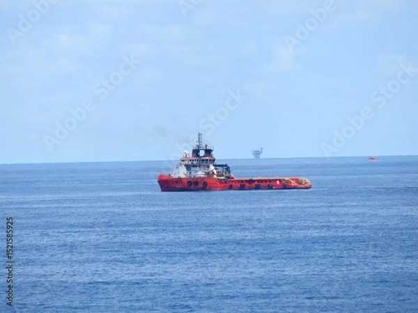Obraz A red supply vessel navigates the vast ocean, a distant oil rig barely visible on the horizon under a pale sky. The calm sea reflects the peaceful atmosphere.