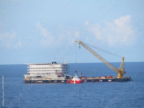 Obraz Offshore platform operation underway, a large crane lifts equipment. A smaller vessel assists nearby. The open ocean under a mostly clear sky provides a dramatic backdrop to this industrial scene.