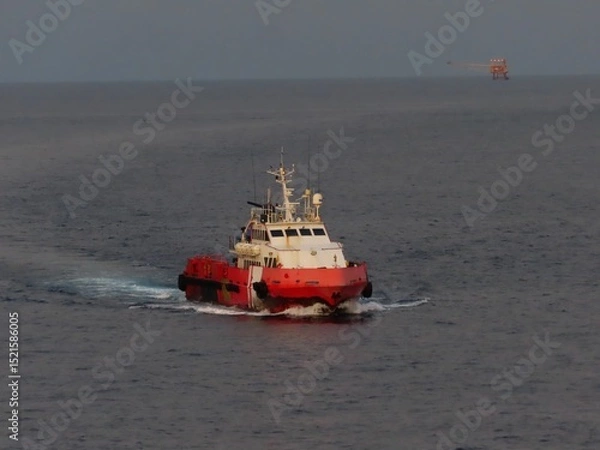 Obraz A vibrant red tugboat cuts through the deep blue sea, leaving a frothy wake. In the distance, an oil rig stands silhouetted against the dusky sky, a solitary sentinel in the vast expanse of water.