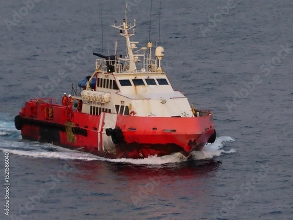 Obraz A powerful red supply vessel cuts through the dark blue ocean waters, its wake a frothy white trail. Crew members are visible on deck, navigating the open sea under a calm, dusky sky.