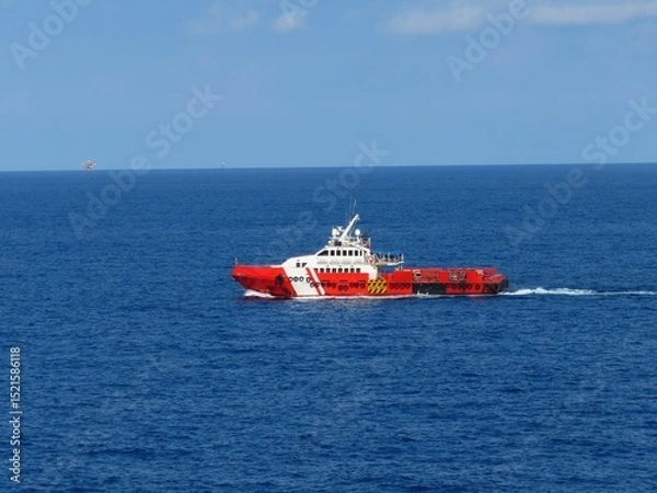 Obraz Red and white vessel navigating the deep blue ocean, under a clear sky. A distant platform is barely visible on the horizon. The ship cuts through the water, leaving a small wake.