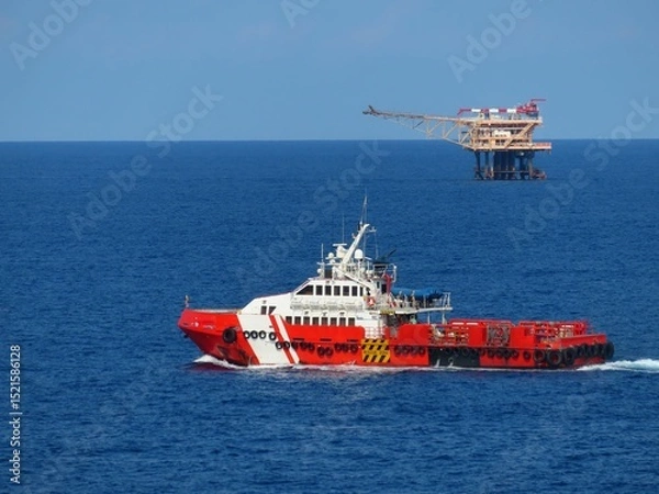 Obraz Red supply vessel navigates the deep blue ocean, approaching a distant offshore oil platform under a clear sky. A scene of maritime industry and vastness.