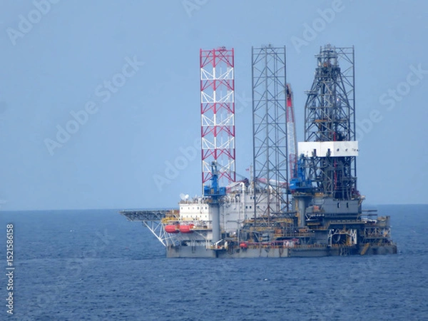 Obraz A large offshore oil rig, composed of multiple metal structures, floats on a calm blue ocean under a clear sky. The rig is the central focus, with the ocean forming a solid background. 