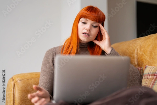 Obraz Woman with red hair sits on a couch, looking stressed while working on a laptop in a modern home setting during daylight hours