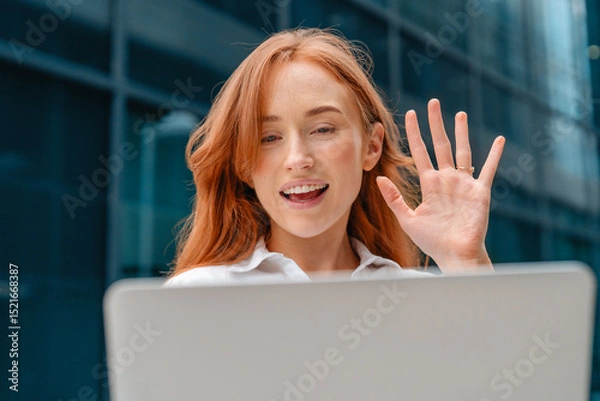 Obraz Young woman waves while video chatting outdoors in a modern urban setting during daylight