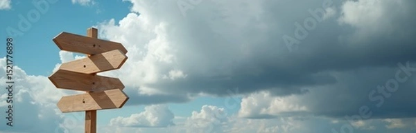 Fototapeta Wooden signpost against sky with clouds. Guidepost with direction arrows show different ways. Symbol of choice, decision making, travel, freedom, exploration, new opportunities crossroads. Ideal for