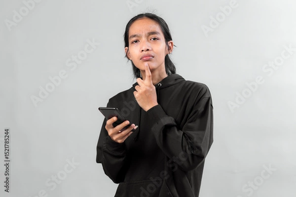 Fototapeta Young Asian man wearing black hoodie holding smartphone with a confused facial expression, touching his chin while looking at camera, isolated on grey background in studio.