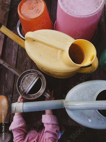 Fototapeta Colorful Gardening Tools and Child's Hands on Wooden Deck – Top View. Rustic gardening scene featuring colorful plastic buckets and watering cans arranged on an aged wooden deck.