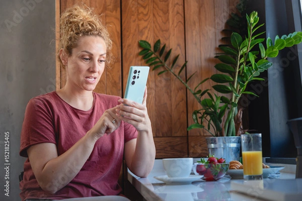 Fototapeta Nervous woman with curly hair looks at smartphone in modern kitchen, sitting by a breakfast table with juice, coffee, and fruit, appearing anxious or uncertain about a message.