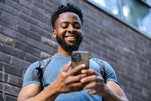 Fototapeta A young man happily interacting with his smartphone, showcasing joy and engagement in an urban setting with a brick wall backdrop.