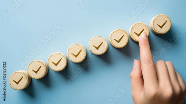Fototapeta Seven wooden tokens with checkmarks increase in size on a blue background as a hand presses the largest, symbolizing progress and achievement in a smooth ascending line
