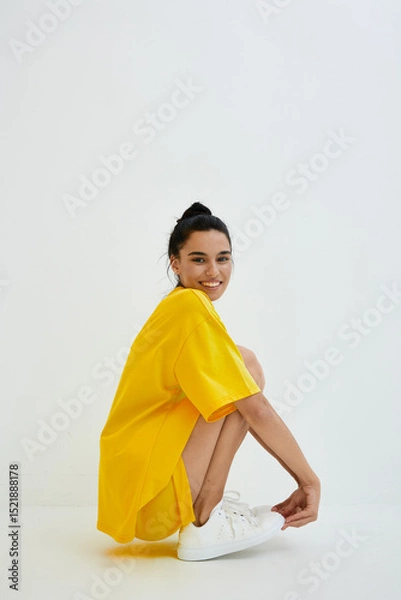 Fototapeta Smiling woman wearing a bright yellow oversized t-shirt and white sneakers, sitting in a relaxed pose against a plain white background with natural lighting.