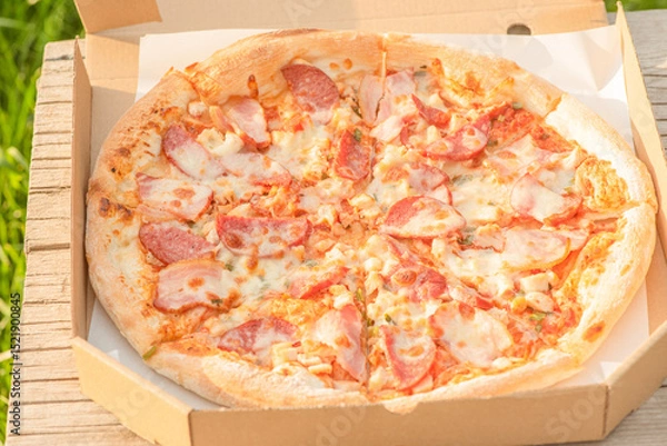 Fototapeta Meat pizza with cheese in a paper box on a wooden board on the grass.