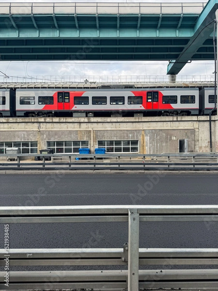Fototapeta A modern commuter train passes under a highway overpass in an urban environment. Symmetrical lines, concrete textures, and transportation infrastructure create a minimalist cityscape.