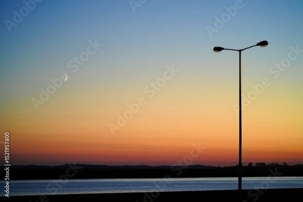 Fototapeta crescent moon over a calm body of water at sunset with a solitary streetlamp silhouetted against a colorful sky