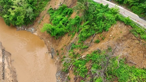 Obraz Drone aerial view shows a landslide on a deforested slope next to a muddy river, illustrating how heavy rains and loss of vegetation accelerate soil erosion and slope failures. Thailand.
