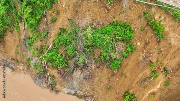 Obraz Drone aerial view shows a landslide on a forested slope beside a muddy river, illustrating how extreme weather and climate change increase soil erosion and slope failures in vulnerable areas.