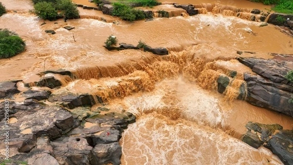Obraz Drone aerial view of sediment-laden floodwaters rushing over rocks, illustrating the severe flooding worsened by deforestation and land mismanagement, highlighting human impact on natural disasters.
