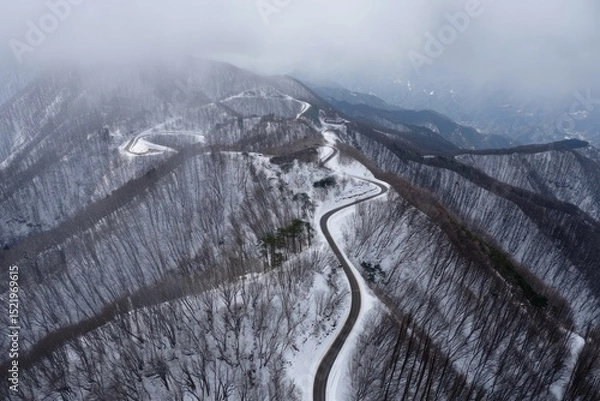 Fototapeta A winding mountain road covered with patches of snow surrounded by leafless trees and foggy mountains under a cloudy sky