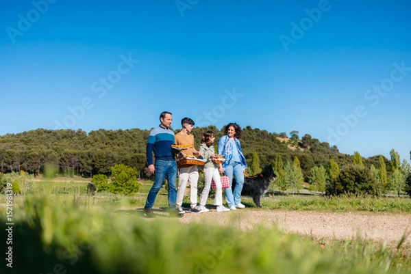 Fototapeta Family walking through a park, carrying picnic baskets and accompanied by their dog, on a beautiful sunny day