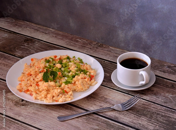 Obraz A plate of egg omelette fried with tomatoes and garnished with a sprig of parsley on a wooden table, next to it a fork and a cup of hot black coffee.