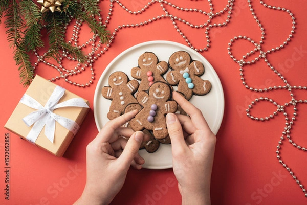 Fototapeta Hands holding gingerbread cookies on a white plate decorated with colorful sprinkles surrounded by holiday decor and gift on a red background. Top view