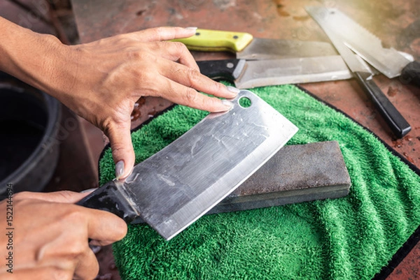 Fototapeta Sharpening knives with grindstone sharpener. Kitchen tools for use. Woman hand sharpens cleaver with whetstone on a green nano cloth pad on old table.